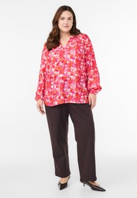 Kwiatowa bluzka w odcieniach różu, czerwieni i bieli, z długimi rękawami zakończonymi elastycznymi mankietami oraz dekoltem v, w zestawie z ciemnobrązowymi spodniami.