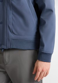 Blaues, glattes Stoffjackett mit geripptem Bund und Bündchen, mit Knopfverschluss und Seitentaschen, kombiniert mit grauen Hosen.