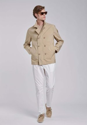 Man draagt een beige dubbelgeknoopt jack, witte broek, bruine suède schoenen en een zonnebril, lopend met handen in de jaszakken.