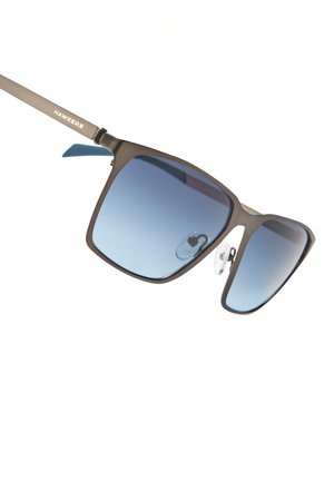 Occhiali da sole con montatura in metallo marrone opaco, lenti quadrate sfumate dal blu scuro al chiaro e naselli regolabili. Logo "HAWKERS" sull'asta.
