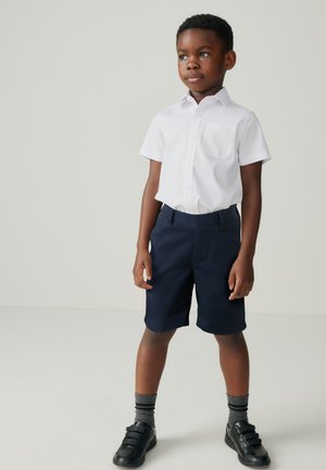Weißes Kurzarmhemd mit einer Brusttasche, kombiniert mit dunkelblauen Shorts. Schwarze Schuhe und graue Socken runden das Outfit ab. Einfaches Design.