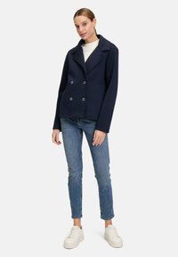 Marineblauer, doppelt knöpfbarer Blazer aus weichem Stoff, mit einem Reverskragen, zwei Fronttaschen und silbernen Knöpfen. Kombiniert mit hellblauen Jeans und weißen Turnschuhen.