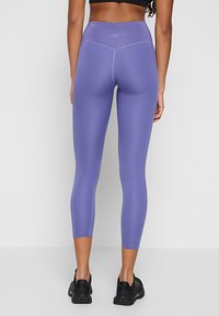 Fioletowe legginsy sportowe z wysokim stanem, gładką fakturą i kontrastowymi detalami szwów. Ubrane z czarnymi butami sportowymi.