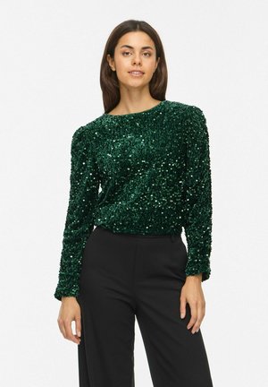 Une femme porte un haut à manches longues vert foncé orné de sequins et un pantalon noir sur un fond blanc uni.