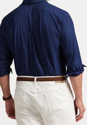 Mann, der ein marineblaues Hemd mit hochgekrempelten Ärmeln und weiße Hose mit braunem Gürtel trägt, von hinten gesehen.