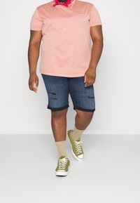 Camiseta rosa claro, pantalones cortos de mezclilla con detalles desgastados y puños enrollados, zapatillas verde oliva, calcetines beige. Conjunto simple y casual.