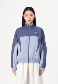Lightweight jacket in navy and light blue, features a high collar, elastic cuffs, and zippered front, with logo detailing on the chest.