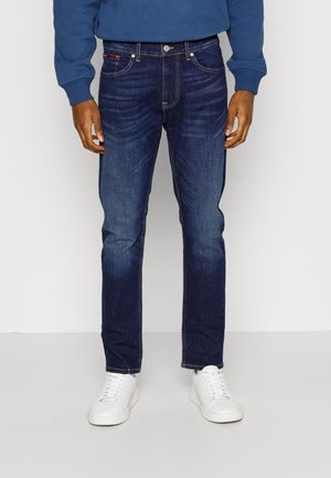 Mann trägt dunkelblaue Jeans, weiße Turnschuhe und ein blaues Langarmshirt, steht vor einem schlichten hellen Hintergrund.