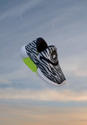 Baskets Nike zébrées noir et blanc avec une semelle verte lumineuse flottant sur un ciel doux bleu et orange.