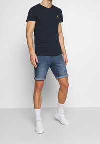 Granatowa bawełniana koszulka z okrągłym dekoltem i żółtym akcentem w postaci logo, połączona z podwiniętymi dżinsowymi szortami, białymi trampkami i białymi skarpetkami.