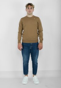 Maglione marrone a coste con scollatura rotonda, abbinato a jeans in denim blu e sneakers bianche. Il maglione presenta un piccolo logo sul petto.