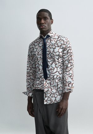 Άνδρας που φοράει λευκό πουκάμισο με μακριά μανίκια με κόκκινες και μαύρες αφηρημένες γραμμές, μπλε γραβάτα και γκρι παντελόνι, στέκεται μπροστά σε ένα απλό γκρι φόντο.