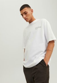 Hvid bomulds t-shirt med korte ærmer, ribbet rund halsudskæring og et lille sort tekstlogo på venstre bryst. Båret med mørke bukser.