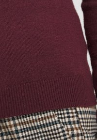 Burgundowa sweter z ribbowym dołem pokazuje gładką teksturę dzianiny, w połączeniu z spodniami w kratę z brązowymi i turkusowymi akcentami.