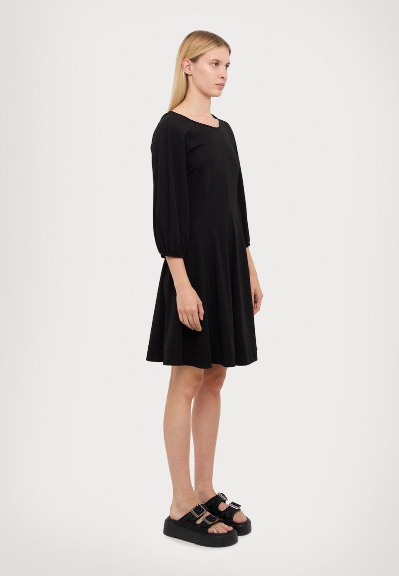 Robe noire mi-longue avec un décolleté arrondi et des manches bouffantes trois-quarts. Le mannequin porte des sandales noires en chunky avec des boucles.