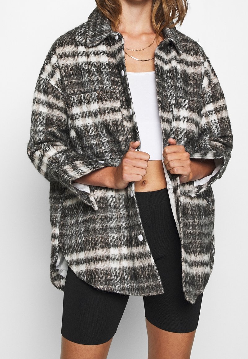 Chemise à carreaux en gris, noir et blanc, en matériau texturé. Dotée d'un col et de boutons, portée sur un haut blanc.