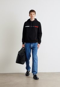 Svart hettegenser med røde og hvite striper over brystet, kombinert med lyseblå jeans og svarte sko, holdende en svart duffelbag.