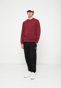 Bordó pulóver kerek nyakkivágással és logó folttal, fekete nadrággal, amely hímzett mintával díszített. A modell fehér tornacipőt visel.