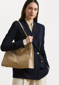 Borsa in suede beige con bordo cucito, manico a catena dorata e un charm decorativo. Il modello indossa un blazer blu navy e una blusa crema.