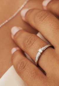 Bague en argent avec une pierre ronde claire au centre, dotée de deux bandes courbées ornées de petites pierres claires sur les côtés. Texture lisse.