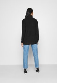 Femme se tenant de dos, portant un pull noir à col roulé, un jean bleu clair et des baskets montantes noires, devant un fond uni.