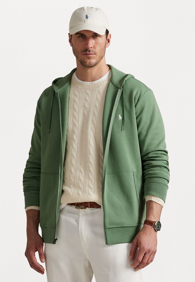 Grön zip-up hoodie med kängurufickor, bärs över en krämfärgad kabelstickad tröja, vit t-shirt och vita byxor. Avslutas med en beige keps.