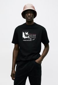 Mann trägt einen hellrosa Anglerhut und ein schwarzes T-Shirt mit einem Jazzmusiker-Motiv und dem Text "Jazz Café Fred Amsterdam City".