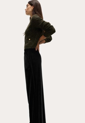 Maglione a maglia testurizzato di colore verde scuro con scollatura rotonda e dettaglio con bottone dorato, abbinato a pantaloni lunghi neri, lisci e aderenti.