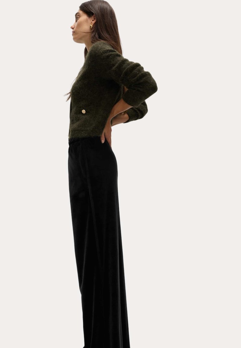 Maglione a maglia testurizzato di colore verde scuro con scollatura rotonda e dettaglio con bottone dorato, abbinato a pantaloni lunghi neri, lisci e aderenti.