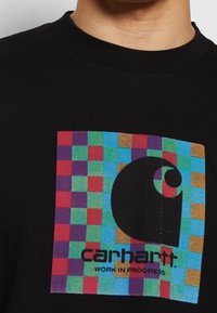 Camiseta de algodón negra con un diseño gráfico a cuadros colorido y un logo negro destacado con el texto "carhartt" y "WORK IN PROGRESS".