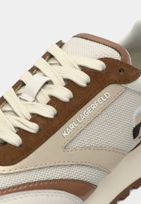 Snøresko med hvit mesh-overdel, brune semskede detaljer, beige overlegg og en synlig merkevarelogo på siden.