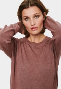 Rjav dolg rokav pulover z rebrasto teksturo in okroglo ovratnikom. Model pozira z rokama za glavo, kar prikazuje mehak material in sproščen kroj.