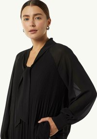 Schwarze plissierte Bluse mit einem gebundenen Kragen, langen Ärmeln und glatter Textur. Verfügt über eine entspannte Passform und seitliche Taschen.