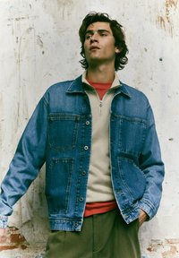 Denim jaka, gaiši zila, ar pogu aizdari priekšā, četriem kabatām, nedaudz pārāk liela piegriezuma, valkāta virs bēša rāvējslēdzēja un sarkanās krekla.