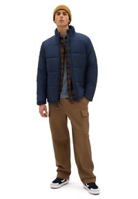 Navy puffer jacket with high collar, layered over a plaid shirt, paired with tan cargo pants and black sneakers, featuring white accents.