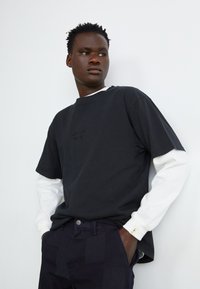 Black cotton t-shirt layered over a white long-sleeve shirt, paired with dark pants. Features a minimal embossed logo on the chest.