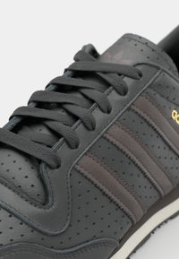 Γκρι δερμάτινο αθλητικό παπούτσι με περφοραρισμένη υφή, καφέ ρίγες και μαύρους κορδόνες. Διαθέτει λευκή σόλα και χρυσές λεπτομέρειες branding.