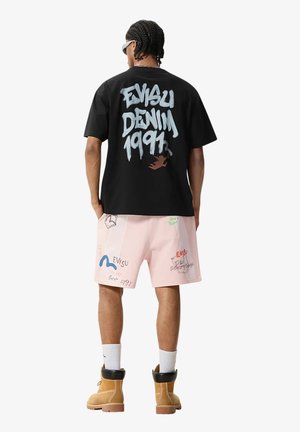 Uomo che indossa una T-shirt nera con scritta grafica "Evisu Denim 1991", pantaloncini rosa con loghi multicolori, calzini bianchi, stivali beige, rivolto di spalle.