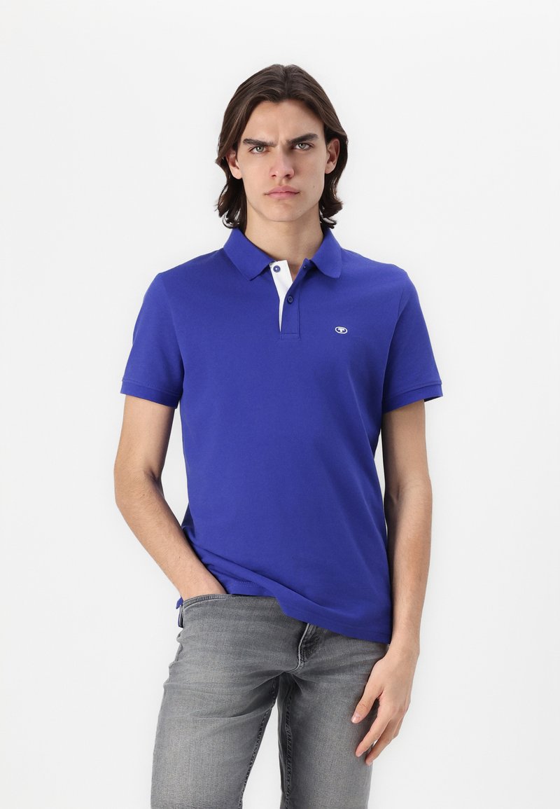 Polo skjorte i lys blå bomuld med hvid krave. Har korte ærmer og et lille logo på venstre bryst. Båret med grå jeans.