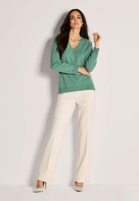 Frau, die an einer Wand steht, trägt einen grünen V-Ausschnitt-Pullover, cremfarbene weite Hosen und gemusterte High-Heels und posiert mit einem erhobenen Arm.