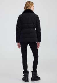 Zwarte pufferjas met een pluizige kraag, voorzien van een doorgestikt ontwerp en lange mouwen. Gecombineerd met zwarte leggings en zwart schoenen met een bontvoering.