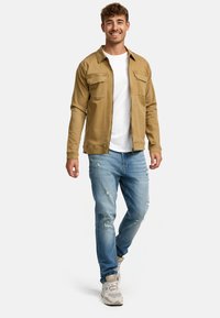 En tanfarvet langærmet skjorte med to brystlommer, båret over en hvid t-shirt. Lyseblå slidte jeans og beige sneakers fuldender outfittet.