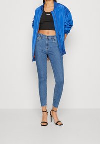 Giacca bombarola blu, top corto nero e jeans skinny azzurri chiari. Sandali neri con tacco a strisce completano l'outfit. Texture morbide, stile casual.