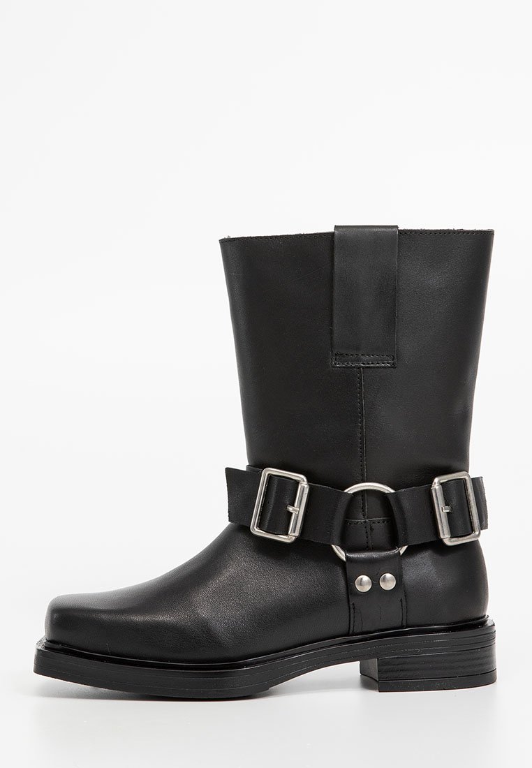 Bottine en cuir noir avec un bout arrondi, hauteur mi-mollet, et une large ceinture avec des boucles et accents argentés. Fini texturé et talon empilable.