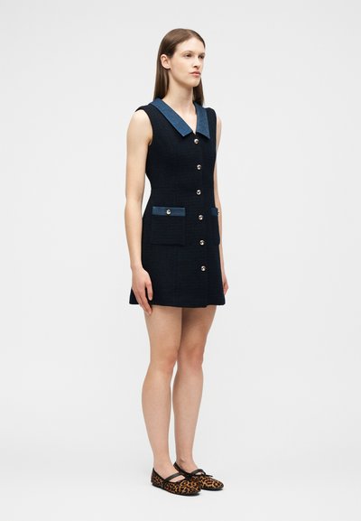 Νεαρή γυναίκα που στέκεται φορώντας ένα αμάνικο μαύρο φόρεμα με μπλε γιακά και λεπτομέρειες στις τσέπες, καθώς και φλατ παπούτσια με λεοπάρ μοτίβο, μπροστά σε απλό ανοιχτό φόντο.