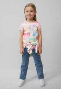 T-shirt met meerkleurige hartpatronen, korte mouwen; een gebonden sweatshirt om de taille; blauwe jeans; witte sneakers. Lichtgrijze achtergrond.