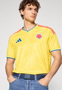 Ung man med lockigt hår som bär en gul colombiansk fotbollströja med röda och blå detaljer, stående med händerna i jeansfickorna.