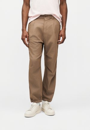 Person iført lysebrune, plisserede bukser med elastiske manchetter og off-white sneakers, stående mod en enkel lys baggrund.