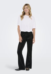 Maglietta bianca oversize abbinata a pantaloni neri in velluto a gamba larga. I pantaloni hanno una texture liscia, mentre le scarpe sono ballerine nere lucide.