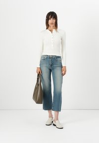 Langermet hvit henley-skjorte med knappestolpe, kombinert med middels vaskede, avkorte jeans og beige slip-on sko; modellen holder en grå veske.